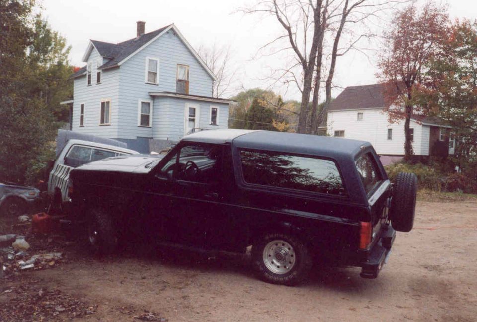 Black Ford Bronco parked in a dirt yard in front of a pale blue two-story house