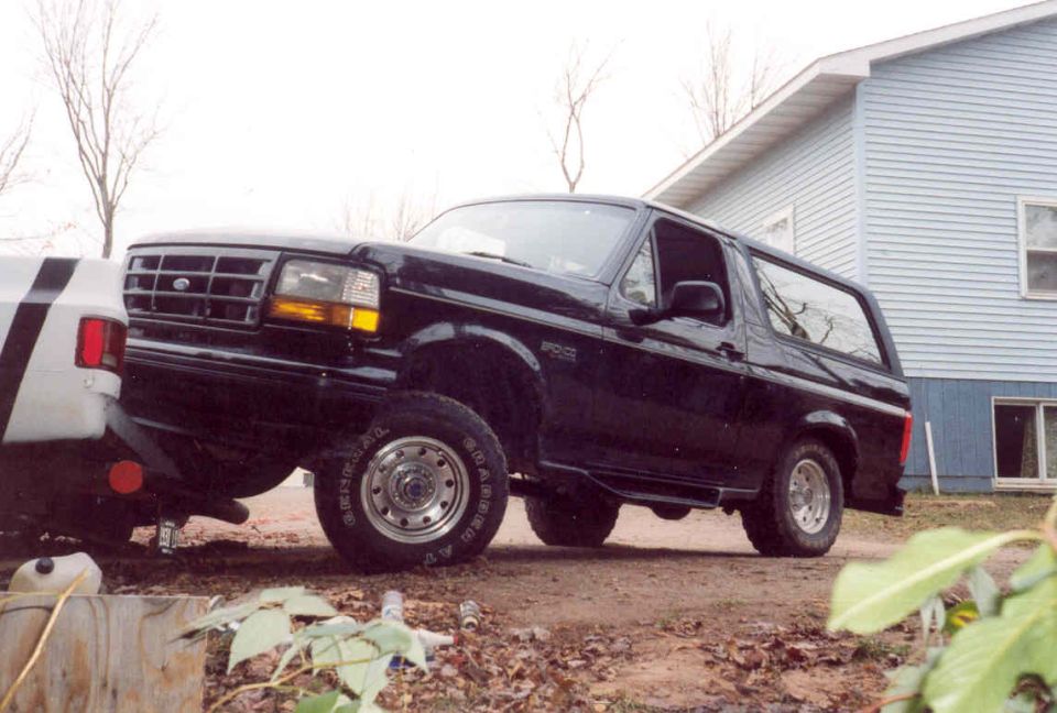 Black Ford Bronco with brush guard parked on gravel beside a blue-sided house