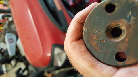 1973 Ford F-100 - A hand holds a metal part with stamped numbers, with a red vehicle in the blurred background.