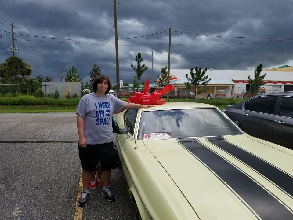 Person in 'I NEED MY SPACE' shirt next to a 1971 Ford Mustang Mach 1 with racing stripes. Red bow on roof