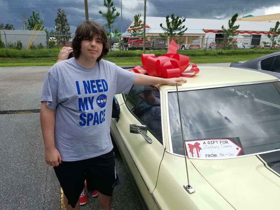 Yellow 1970s coupe with a large red bow on the roof, seen from the front passenger side