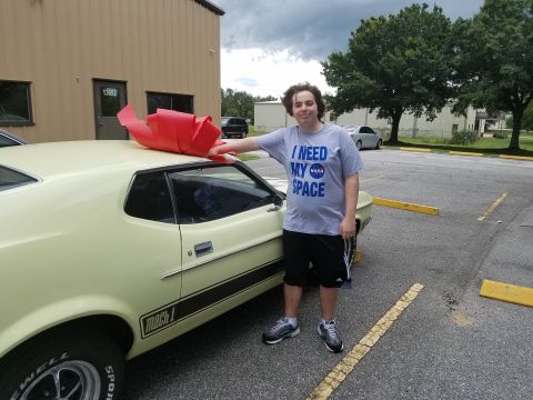 1971 Ford Mustang - Yellow 1971 Ford Mustang Mach 1 with a large red bow on the roof, a man stands beside it.