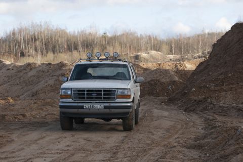 1993 Ford Bronco - A white Ford Bronco drives through a dirt quarry with trees in the background.