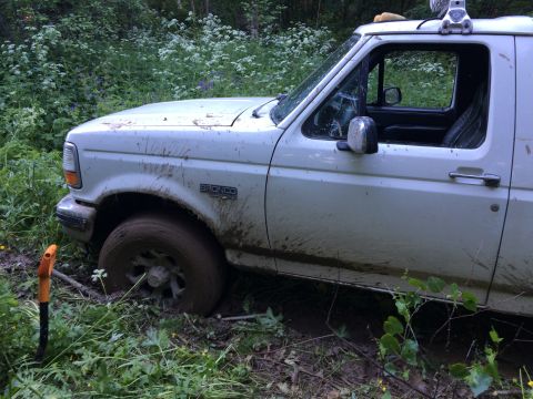 1993 Ford Bronco - A white 1993 Ford Bronco is stuck in the mud, with its front wheel submerged and the windshield cracked.