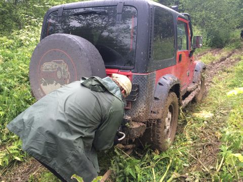 1993 Ford Bronco - A red Jeep Wrangler is stuck in the mud on a trail, with a person attempting to attach a tow strap.