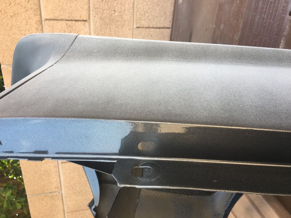 Close-up of a metallic gray car part, possibly a fender or bumper section, showing mounting holes