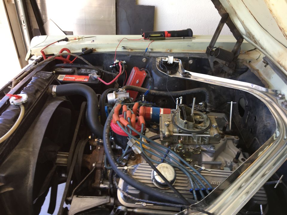 Open engine bay of a classic car showing V8 with four-barrel carburetor, finned valve covers, and red plug wires