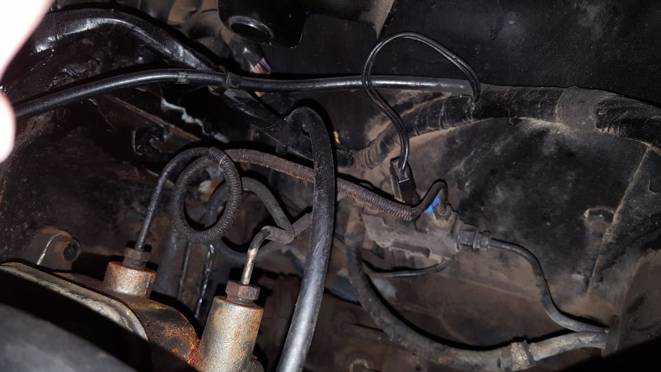 Brake lines and master cylinder in a dark, possibly engine bay area. Rusty metal components and black hoses are visible