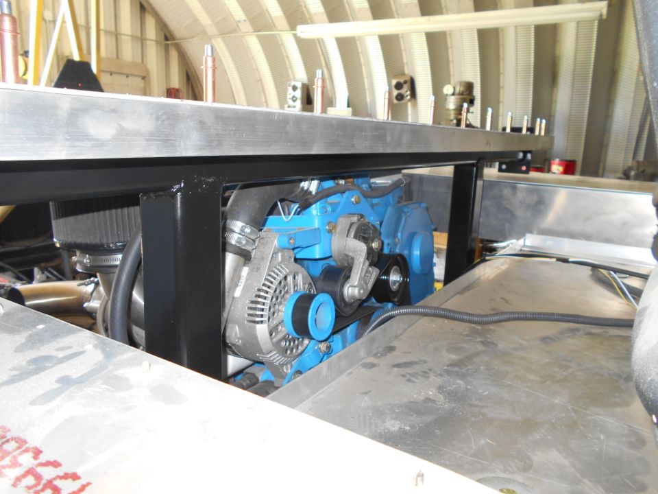 Close-up of a blue engine block with an alternator, belts, and air intake. Some metal sheeting is visible in the foreground