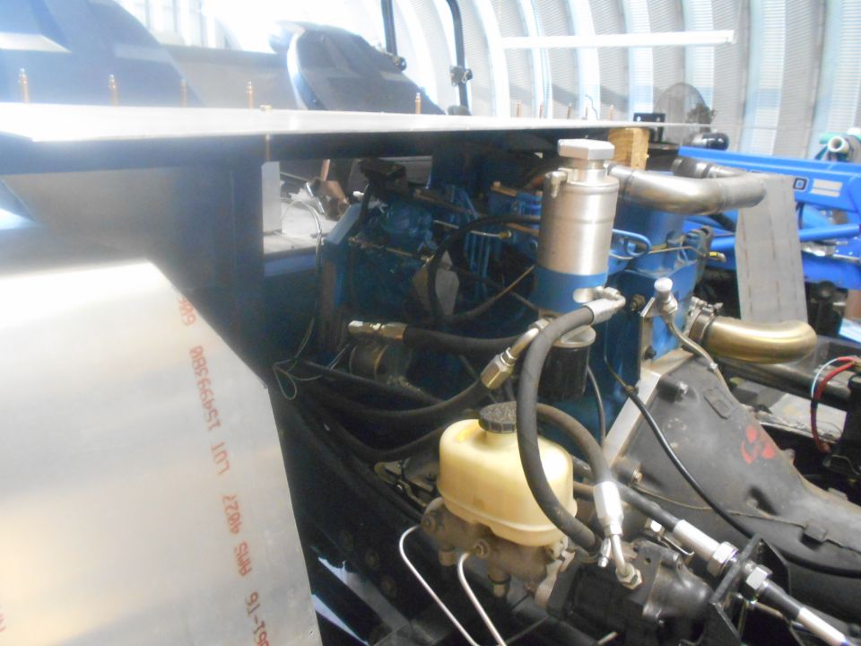 Engine bay with blue engine block, hoses, and brake fluid reservoir. Some metal sheeting is in the foreground