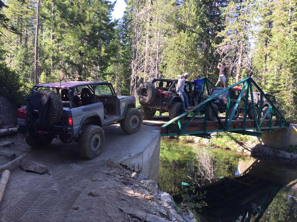 Gray 1979 Ford Bronco with roll cage crossing a bridge with other off-road vehicles