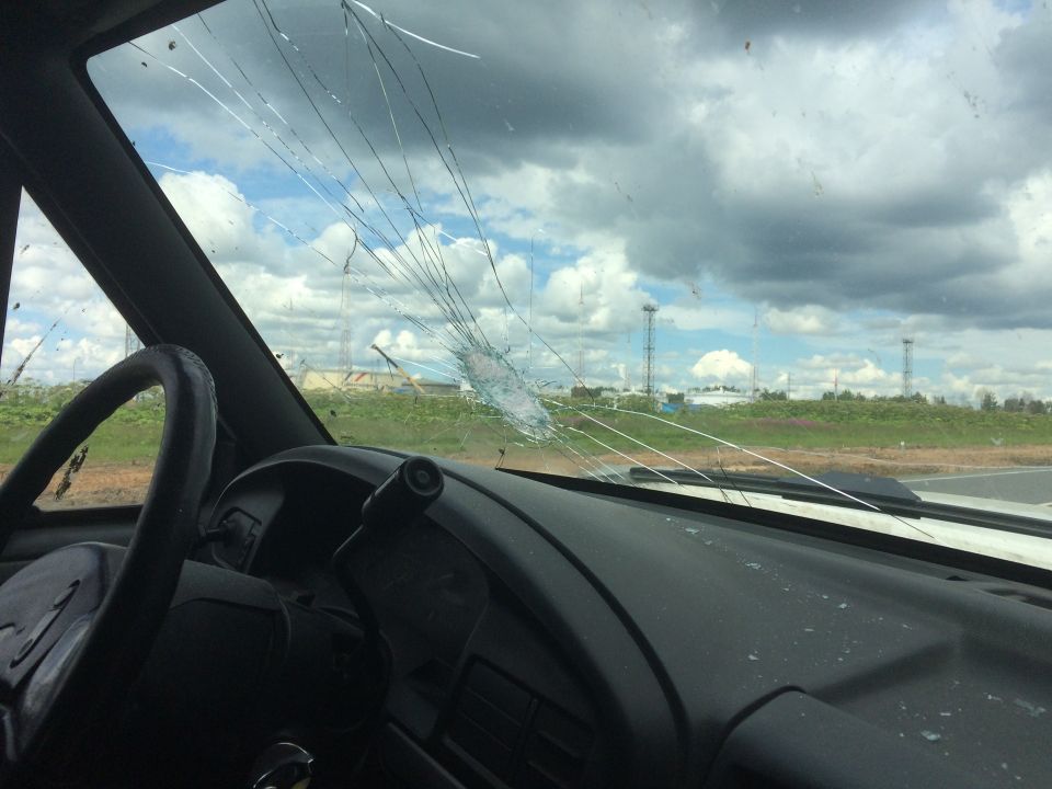 View from inside a vehicle looking out a cracked windshield at a cloudy sky and distant structures