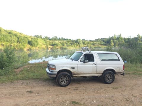 1993 Ford Bronco - White 1993 Ford Bronco sits parked on a dirt path beside a calm lake with wooded hills beyond.