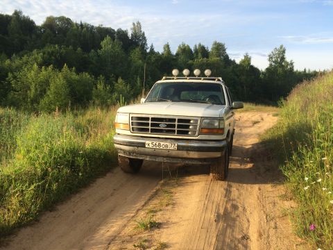1993 Ford Bronco - A white Ford Bronco drives down a dirt road surrounded by trees.