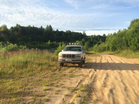 1993 Ford Bronco - A white 1993 Ford Bronco is parked on a dirt road surrounded by trees and tall grass under a cloudy sky.