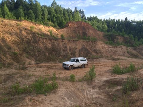 1993 Ford Bronco - A white 1993 Ford Bronco navigates a sandy, off-road terrain with wooded hills in the background.