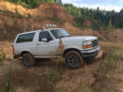 1993 Ford Bronco - White Ford Bronco navigates a muddy off-road course with a wooded hillside in the background.