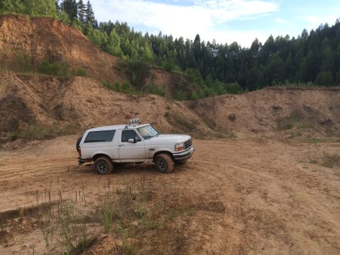 1993 Ford Bronco - White 1993 Ford Bronco navigates a sandy off-road terrain with wooded hills under a cloudy sky.