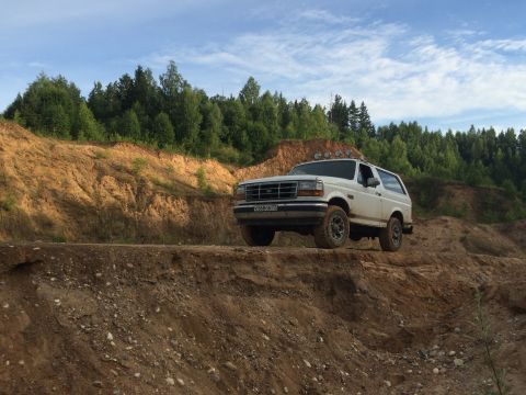1993 Ford Bronco - A white Ford Bronco drives on a dirt path, surrounded by trees under a cloudy sky.