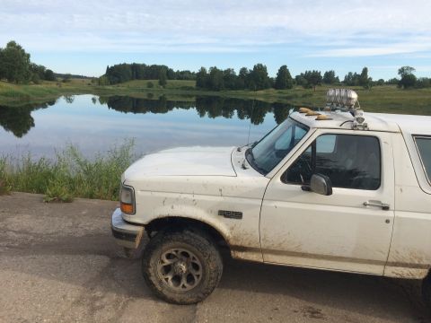 1993 Ford Bronco - A white Ford Bronco is parked on a dirt road next to a calm lake under a cloudy sky.