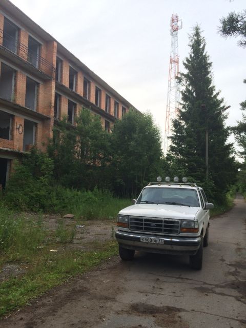 1993 Ford Bronco - A white SUV is parked on a dirt road in front of an abandoned building with a cell tower in the background.