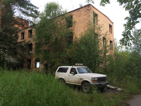 1993 Ford Bronco - A white 1995 Ford Bronco sits abandoned next to a derelict brick structure, surrounded by tall grass and trees.