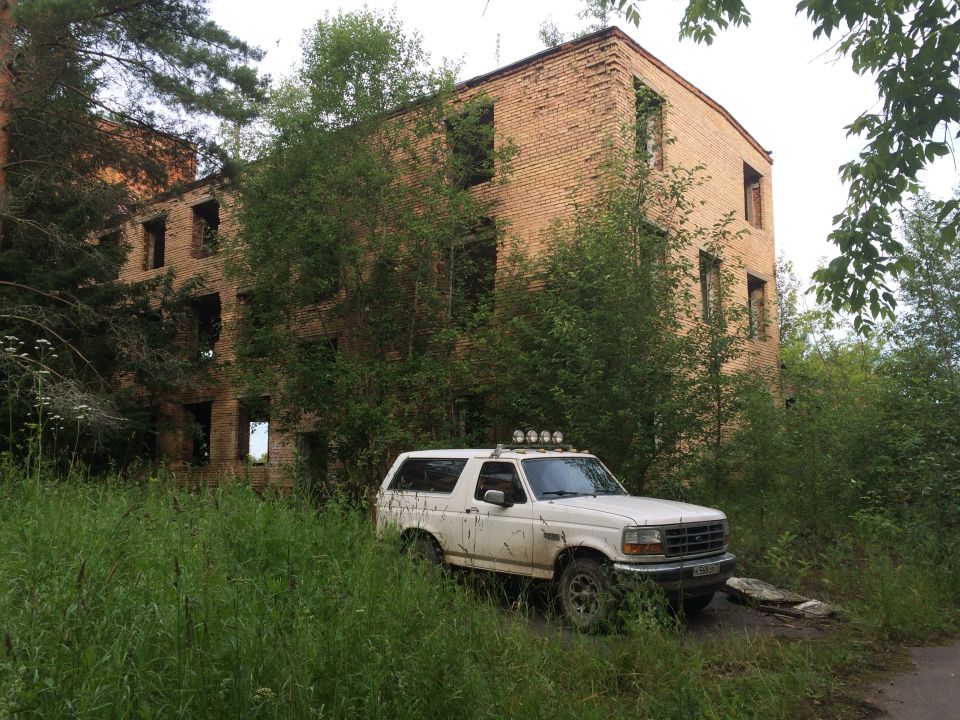 White 1995 Ford Bronco with roof lights parked in overgrown grass near an abandoned building