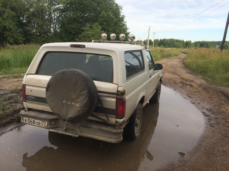 White Ford Bronco with spare tire on rear, parked in a muddy puddle on a dirt road