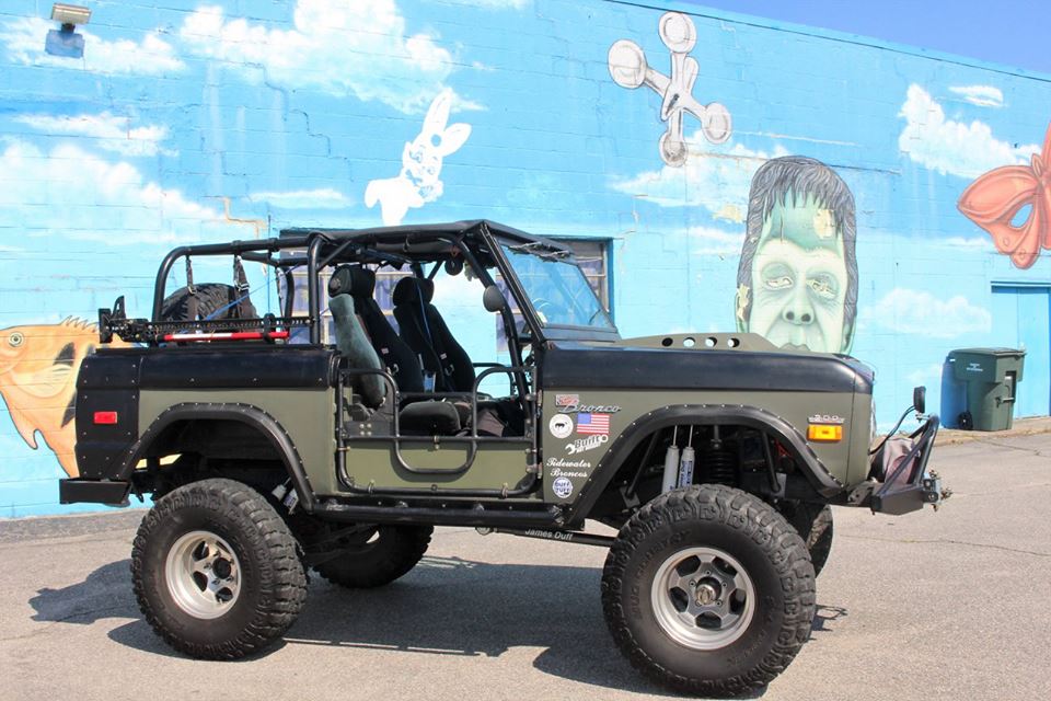 Olive green and black Ford Bronco with a roll cage and large tires, parked in front of a mural