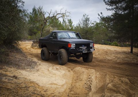 1989 Ford Bronco - Modified black Ford Bronco navigates a sandy off-road trail under an overcast sky.