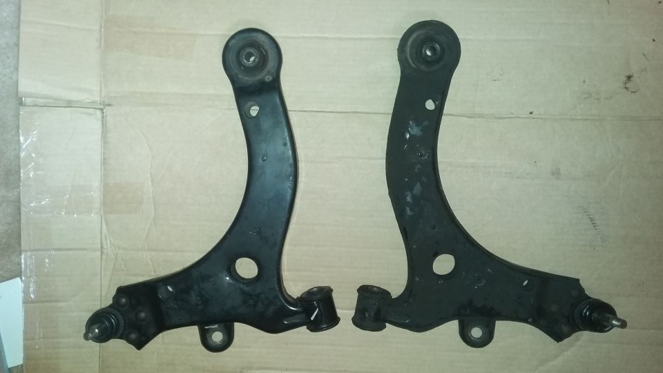 Two black car control arms are laid out on a cardboard surface. A watermark reads "SUPERMOTORS www.supermotors.net"