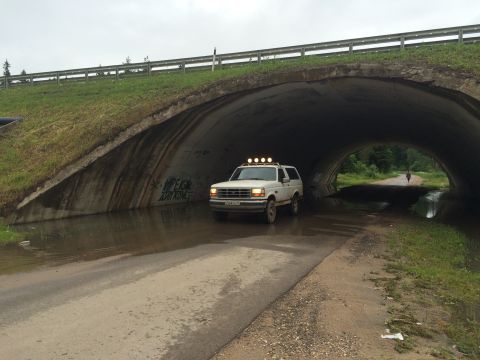 1993 Ford Bronco - A white 1993 Ford Bronco drives through a flooded underpass on a cloudy day. The vehicle has four auxiliary lights mounted on its roof.