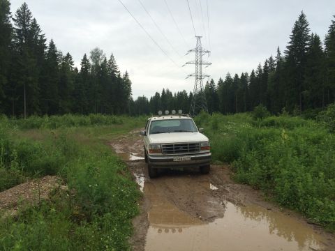 1993 Ford Bronco - A white 1993 Ford Bronco is parked on a muddy dirt road in a forest clearing, with a power line tower visible in the background.