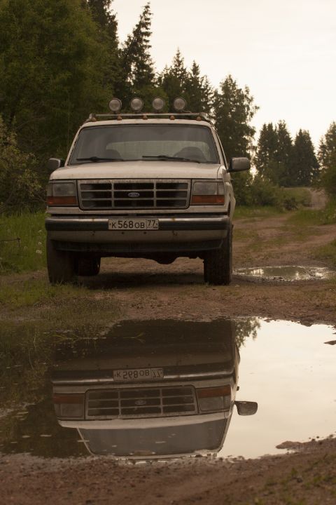 1993 Ford Bronco - A white Ford Bronco is parked on a dirt road with puddles reflecting the vehicle. The front of the SUV is visible, featuring a roof rack wi…