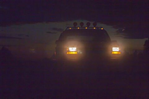 1993 Ford Bronco - The front of a dark-colored SUV is visible at night, illuminated by its headlights and four auxiliary lights mounted on the roof rack. The…