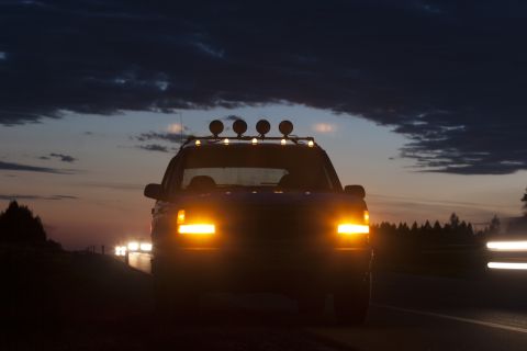 1993 Ford Bronco - A dark blue SUV with its headlights and roof-mounted lights illuminated, driving on a road at dusk. The vehicle is seen from the front, wit…