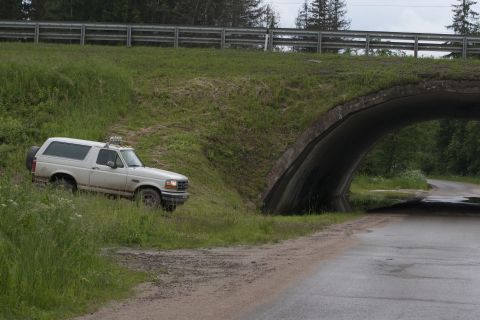 1993 Ford Bronco - A white 1990s Ford Bronco is parked on a grassy incline next to a concrete underpass. The vehicle has off-road lights mounted on the roof r…