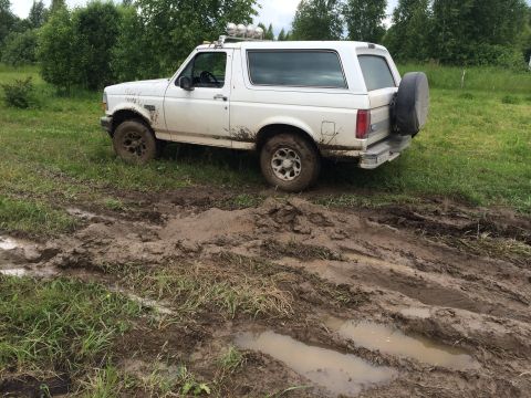 1993 Ford Bronco - A white 1990s Ford Bronco is parked on a muddy track with puddles and tire ruts. The vehicle is covered in mud and has a spare tire mounted…