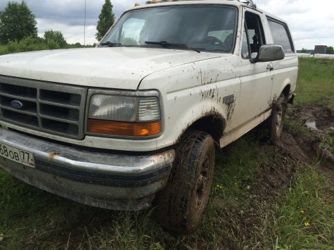 1993 Ford Bronco - A white Ford Bronco is shown from the front 3/4 angle, covered in mud after driving off-road. The vehicle is parked in a grassy field with…