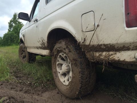 1993 Ford Bronco - A white Ford Bronco is shown from a low angle, heavily covered in mud after driving through a muddy field. The rear tire and fender are par…