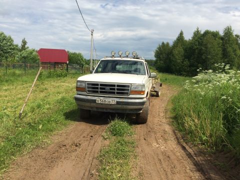1993 Ford Bronco - A white Ford Bronco with a trailer attached is parked on a muddy dirt road in a rural setting with trees and a red-roofed building in the b…