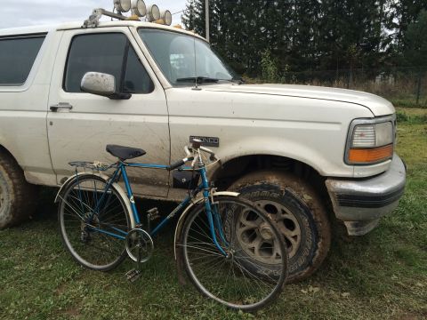 1993 Ford Bronco - A muddy white 1990s Ford Bronco is parked next to a blue bicycle on a grassy area. The Bronco has off-road lights mounted on the roof rack…