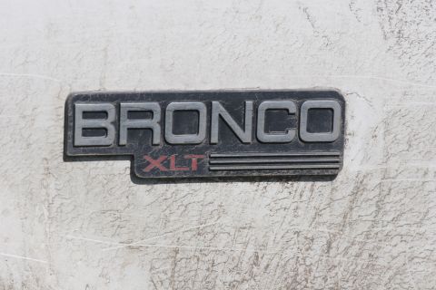 1993 Ford Bronco - A close-up view of a Bronco XLT badge on a white vehicle. The badge is black with silver lettering for BRONCO and red lettering for XLT.