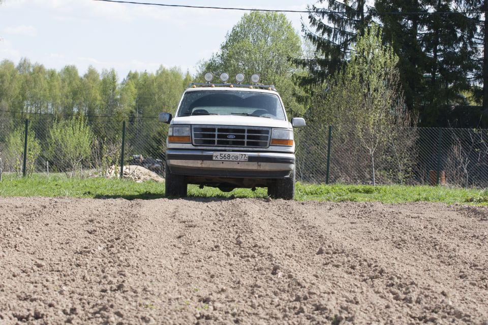 White Ford Bronco with roof lights parked on dirt in front of fence and trees.