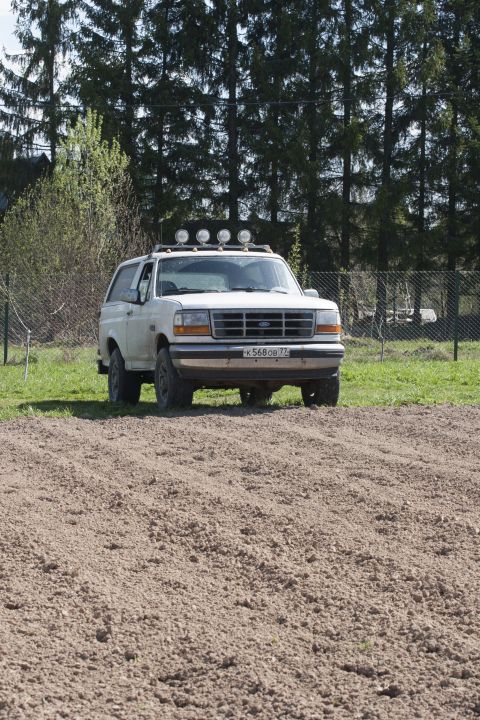 1993 Ford Bronco - A white 1993 Ford Bronco is parked on a dirt patch in front of a grassy area and a line of trees. The SUV has four auxiliary lights mounted…
