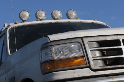1993 Ford Bronco - Close-up front view of a dusty white 1990s Ford Bronco with four round auxiliary lights mounted on the roof. The grille and headlight assem…