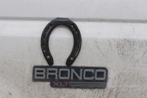 1993 Ford Bronco - A black horseshoe emblem is mounted above a black and silver "BRONCO XLT" badge on a white vehicle. The vehicle appears to be a 1993 Ford B…
