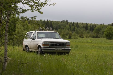1993 Ford Bronco - A white, two-door Ford Bronco is parked in a grassy field with a forest in the background under an overcast sky. The vehicle has auxiliary…