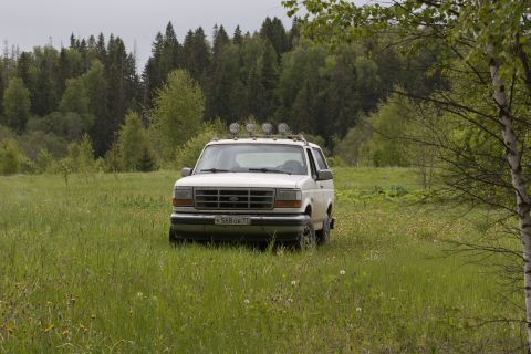 1993 Ford Bronco - A white Ford Bronco is parked in a grassy field with a forest in the background. The vehicle has four auxiliary lights mounted on its roof…