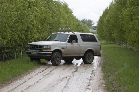 1993 Ford Bronco - A white Ford Bronco is parked on a muddy dirt road lined with trees. The vehicle has four auxiliary lights mounted on the roof rack.